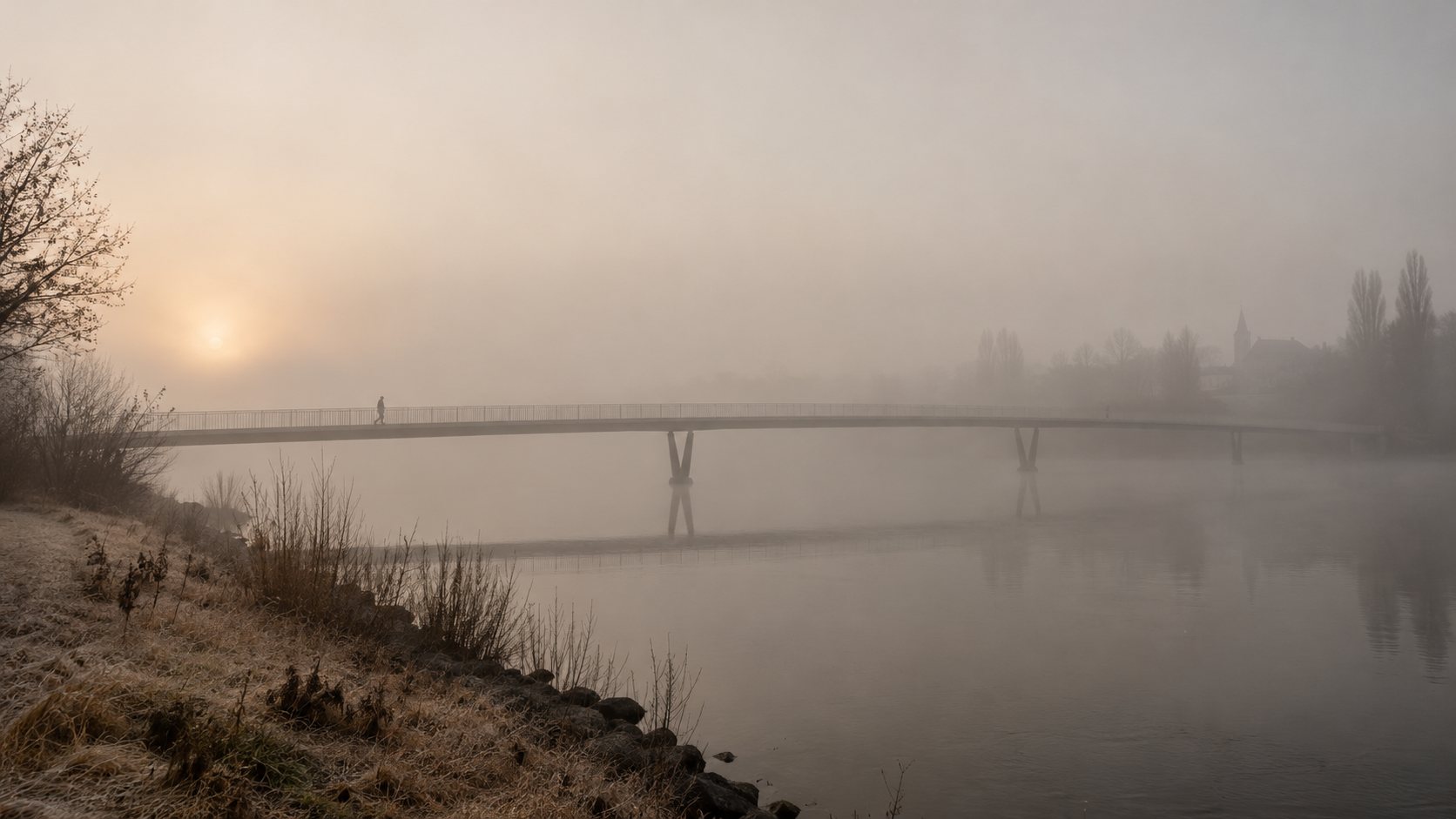 A long footbridge connecting two banks of a wide river in fog.